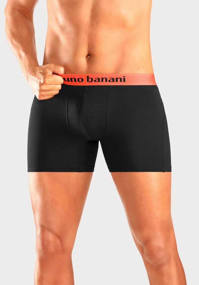 Männlicher Oberkörper und Beine mit schwarzen Boxer-Shorts mit einem orangefarbenen Bund, auf dem „bruno banani“ steht, Hand hält den Bund.