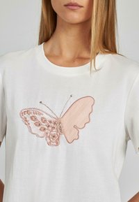 Camiseta de algodón blanca con un motivo de mariposa bordado en rosa, con detalles intrincados y acentos de cuentas en las alas.
