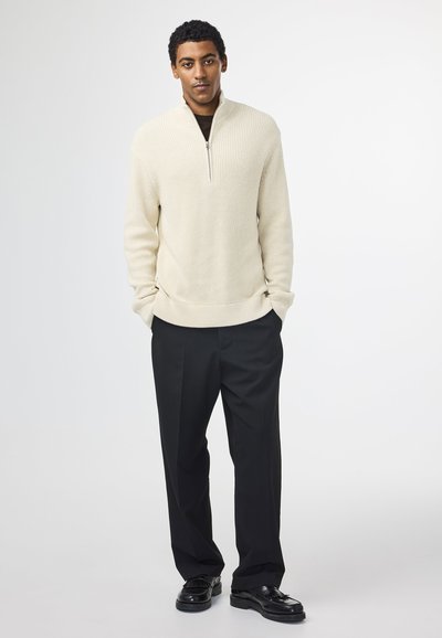 Beige ribbed striktrøje med lynlås i kraven, parret med sorte dressbukser og sorte sko. Modellen står foran en neutral baggrund.