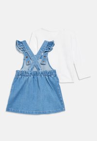 Hvit langermet skjorte parret med en blå denim snekkerkjole med volanger, kryssede skulderstropper og elastisk midje.