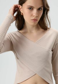 Beige top met lange mouwen, een v-hals en een geribbelde textuur, met een overslagontwerp en zichtbare stiksel details.