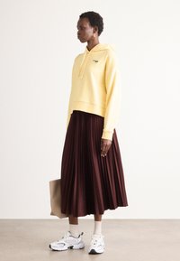 Felpa cropped gialla con logo verde, abbinata a una lunga gonna plissettata bordeaux. Sneakers bianche e borsa beige completano il look.