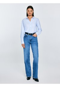 Sieviete stāv, valkājot gaiši zilu pogājamu kreklu, augsta vidukļa zilas džinsas, melnu jostu un melnas smailas kurpes uz vienkārša fona.