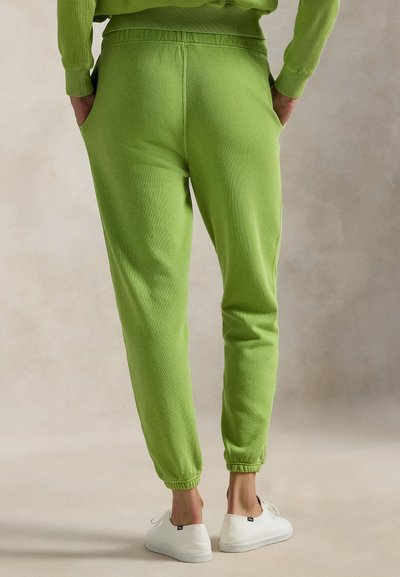 Πράσινη φόρμα από μαλακό ύφασμα, με ελαστική μέση και μανσέτες, πλαϊνές τσέπες και άνετη εφαρμογή. Φορεμένη με λευκά παπούτσια.