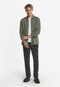 Grön knäppskjorta över en vit t-shirt, kombinerad med svarta byxor och svarta skor. Skjortan har långa ärmar och en avslappnad passform.