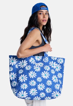 Niebieska torba na ramię w kwiatowy wzór, wykonana z wytrzymałej tkaniny, z białymi stokrotkami z turkusowymi środkami, dużymi uchwytami i naszywką z logo.