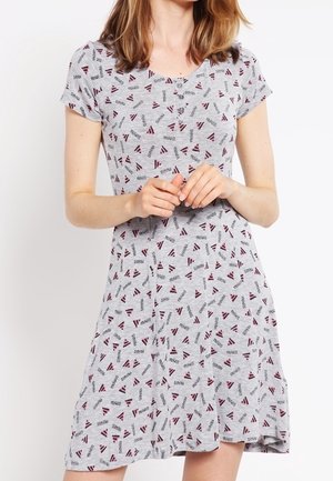 Frau trägt ein graues, knielanges Kleid mit kurzen Ärmeln und kleinen dreieckigen Mustern in Bordeauxrot und Schwarz, steht mit gefalteten Händen.