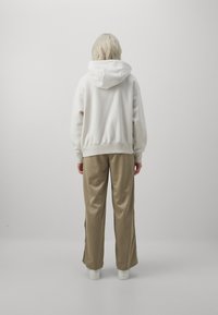 Bílá fleecová mikina s velkou kapucí, spárovaná s béžovými kalhotami s širokými nohavicemi. Kalhoty mají boční rozparky a jsou vyrobeny z hladkého materiálu.