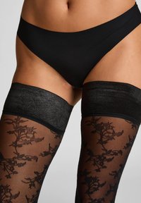 Nahaufnahme einer Person, die schwarze florale Spitzenstrümpfe bis zum Oberschenkel und nahtlose schwarze Unterwäsche vor einem schlichten Hintergrund trägt.