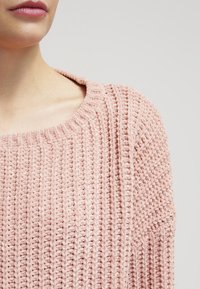 Texturovaný, světle růžový pletený svetr s širokým výstřihem. Má žebrovaný vzor a měkký, nadýchaný materiál. Volný, uvolněný střih.