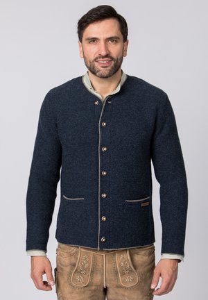 Mann trägt dunkelblaue, geknöpfte Wolljacke und beige traditionelle bestickte Lederhose, steht vor einfarbigem Hintergrund.