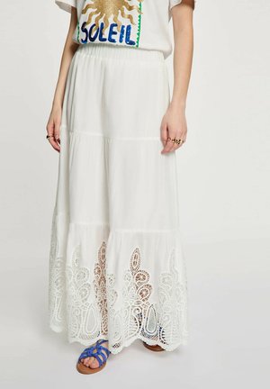 Donna che indossa una gonna lunga bianca a balze con orlo in pizzo, sandali blu con cinturini e T-shirt bianca con sole dorato e scritta "SOLEIL".