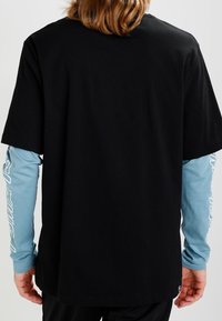 Schwarzes kurzärmliges T-Shirt, das über einem hellblauen langärmeligen Hemd getragen wird. Es hat einen glatten Stoff und einen lockeren Schnitt, mit sichtbaren Nähten und Bündchen.