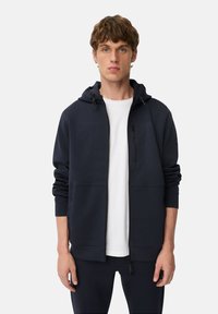 Marineblå zip-up hoodie med en frontlomme, justerbar hætte og tekstureret stof. Bæres over en hvid t-shirt med minimal branding.