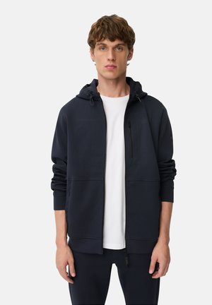 Sweat à capuche zippé navy avec une poche avant, capuche réglable et tissu texturé. Porté par-dessus un t-shirt blanc, avec un branding minimal.