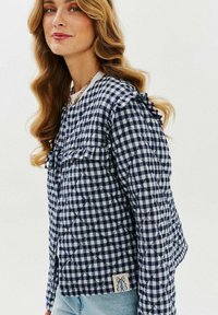 Pikowana granatowa i biała kurtka w kratę w stylu gingham z falbanami na ramionach, z małą naszywką z logo u dołu. Gładki materiał z zabawnym wzorem.