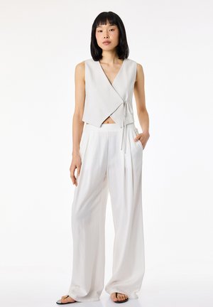 Vrouw met zwart haar, gekleed in een mouwloos lichtgrijs wikkeltop en wijde witte broek, staand met één hand in de zak tegen een witte achtergrond.