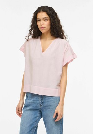 Kobieta o długich kręconych włosach, ubrana w luźny, jasnoróżowy top z krótkim rękawem i dekoltem w serek oraz niebieskie dżinsy, stojąca na tle białego tła.