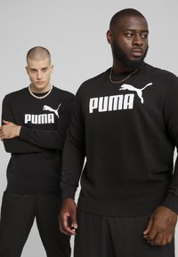 Čierna mikina s veľkým bielym logom "PUMA" a dizajnom skákajúcej pumy. Hladká látka, dlhé rukávy a rebrované manžety.