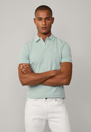 Svetlozelená polo košeľa s klasickým golierom, krátkymi rukávmi a malým vyšitým logom. Na sebe má biele džínsy, model má prekrížené paže.