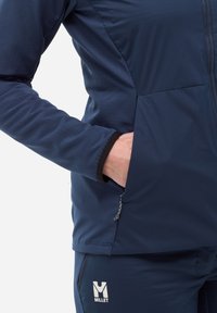 Veste de sport bleu marine au design ajusté, en tissu texturé, avec une poche zippée. Comprend un poignet noir contrastant et un logo.