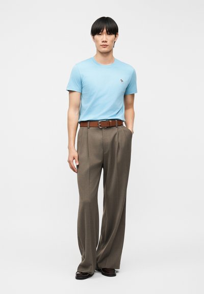 Νεαρός άνδρας στέκεται φορώντας ανοιχτό μπλε κοντομάνικο πουκάμισο, καφέ ζώνη, φαρδιά παντελόνια σε απόχρωση ταουπ και σκούρα καφέ παπούτσια, μπροστά σε απλό φόντο.