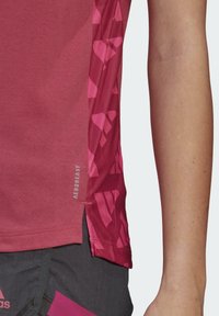 Czerwona sportowa koszulka z krótkimi rękawami, z teksturowanym różowym geometrycznym wzorem wzdłuż boku oraz etykietą "AEROREADY" blisko dołu.