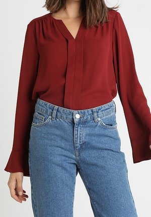 Frau trägt eine dunkelrote Bluse mit langen Ärmeln, in eine hoch taillierte blaue Jeans gesteckt, steht vor einem schlichten Hintergrund.