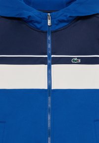 Blå zip-up huvtröja med huva, med marinblå, vit och ljusblå horisontella ränder. Lacoste-logga i grönt till vänster.