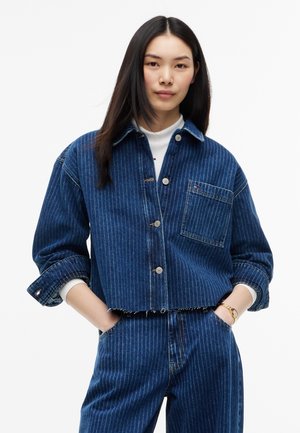 Ung kvinde med langt mørkt hår iført blå denimjakke med striber og matchende bukser, hænderne i lommerne, står mod en hvid baggrund.