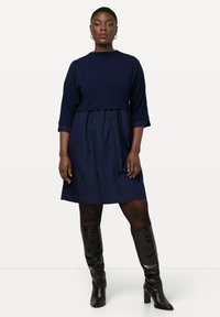Marineblaues Kleid mit Dreiviertelärmeln, tailliertem Oberteil und lockerem Rock. Getragen mit kniehohen schwarzen Stiefeln, glatter Textur.