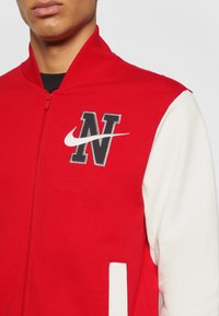 Homme portant une veste rouge et blanche avec la lettre N noire et le logo Nike swoosh blanc sur la poitrine, zippée.