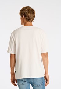Camiseta de algodón blanca con mangas cortas y un cuello redondo clásico. El dobladillo tiene una ligera curva y lleva una pequeña etiqueta de la marca en el lateral.