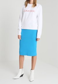Sweatshirt blanc avec le logo "Calvin Klein" en rose, associé à une jupe midi côtelée bleu et des bottes ankle blanches brillantes à talons.