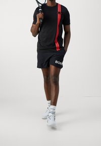 Camiseta atlética negra con rayas verticales rojas y logo, combinada con pantalones cortos negros que llevan "BORGO" en blanco. Zapatillas y calcetines blancos.