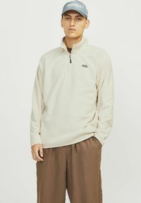 Beige fleece-tröja med kvartslång dragkedja och svart logotyp på bröstet, ihop med lösa bruna byxor. Mjuk textur och avslappnad passform.