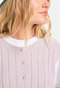 Gilet tricoté rose clair avec une texture en côtes, comportant quatre boutons assortis. Porté par-dessus un t-shirt blanc à encolure ronde.