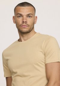 T-shirt beige à manches courtes en tissu doux et lisse, avec un col rond et des détails de couture subtils.