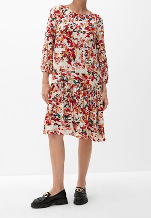 Femme portant une robe fleurie ample arrivant au genou avec des manches trois-quarts et des mocassins noirs ornés d'une chaîne dorée.