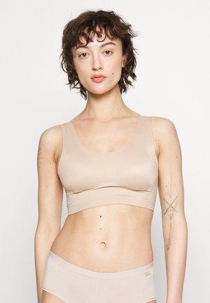 Mujer con cabello corto y rizado, vistiendo un sujetador deportivo beige y ropa interior, de pie contra un fondo claro y liso, con tatuajes visibles en los brazos.