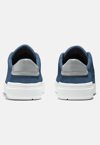 Zapatillas de ante azul con acentos en el talón gris, suelas de goma blanca y plantillas texturizadas; diseño minimalista sin cordones visibles.