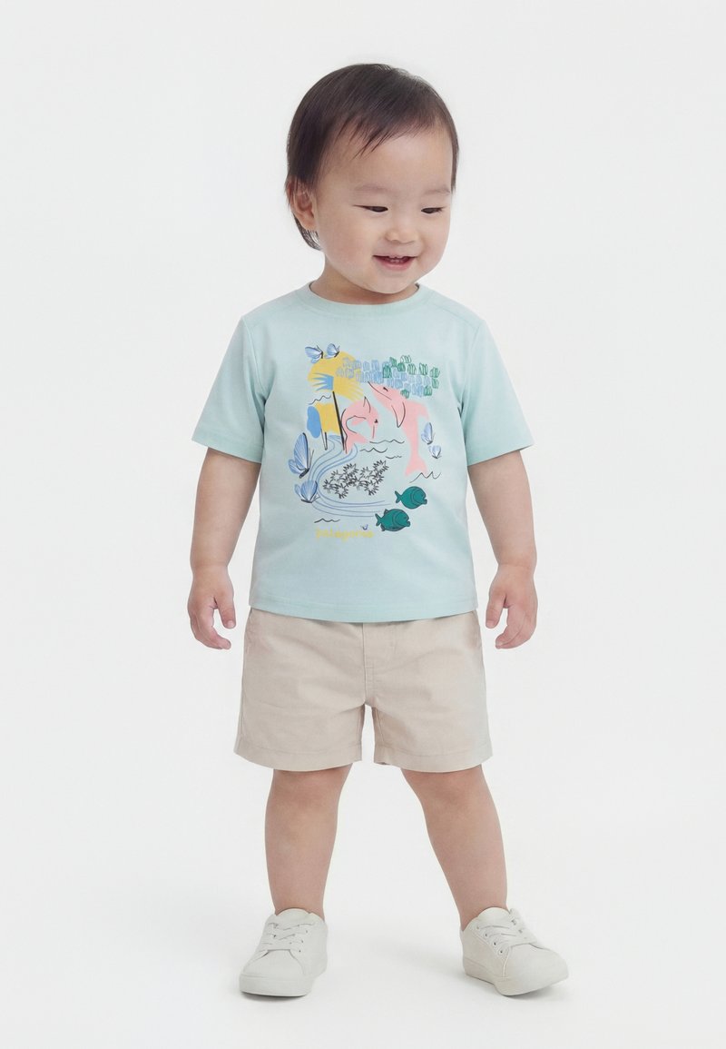 Νήπιο με ανοιχτό μπλε φανελάκι με πολύχρωμα θαλάσσια ζώα, μπεζ σορτς και λευκά αθλητικά παπούτσια, στέκεται και χαμογελά σε λευκό φόντο.