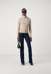 Pull à col roulé beige à côtes avec des manches longues, associé à un jean évasé bleu foncé et des escarpins noirs à bout pointu. Petit sac à main noir.