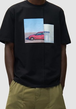 Persona che indossa una t-shirt nera con grafica di un'auto rossa accanto a un muro bianco con un cartello "Antwerpen", abbinata a pantaloni beige.