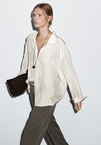 Silk cream button-up shirt with a relaxed fit, paired with olive trousers. Model holds a dark brown suede clutch. Clean background.
