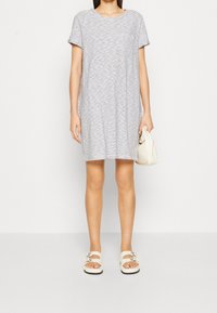 Grå och vit randig t-shirtklänning med korta ärmar och bröstficka, kombinerad med vita sandaler. Tyget ser mjukt och avslappnat ut.