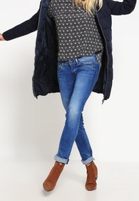 Vadderad svart kappa över en mönstrad svart och vit topp, kombinerad med blå jeans och bruna mocka ankelskorn med uppvikta fållar.