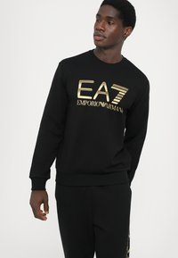 Svart hettegenser laget av bomull med en gyllen "EA7 Emporio Armani" logo på fronten. Har rund hals og ribbestrikkede mansjetter og kant.