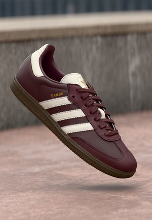 Baskets Adidas Samba bordeaux avec rayures blanches et semelle en gomme, positionnées en l'air contre un sol en béton rosé et un mur en pierre en arrière-plan.