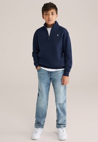 Mornarsko modra zip-up kapuca s majhnim logotipom, pod njo bela majica s kratkimi rokavi, v kombinaciji s svetlo modrimi raztrganimi džinsi in belimi tenisicami.